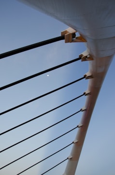 A close-up view of the structural elements of a bridge or similar architecture, featuring long black support cables attached to a large white beam, with a clear blue sky in the background.