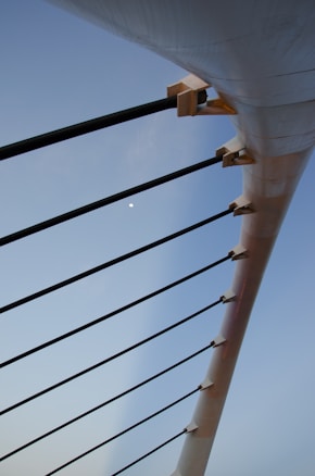 A close-up view of the structural elements of a bridge or similar architecture, featuring long black support cables attached to a large white beam, with a clear blue sky in the background.