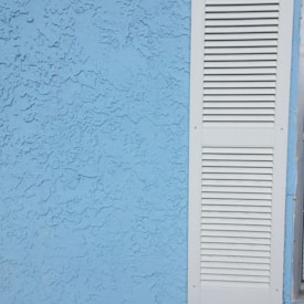 A textured blue stucco wall is situated next to a white louvered shutter.