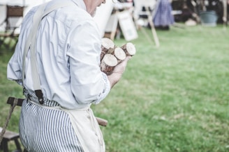 A person wearing a light blue shirt and striped apron is holding several cut sections of wood. The background is a grassy area with some blurred wooden structures, suggesting a rustic or outdoor setting.