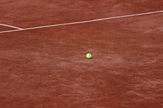 A tennis ball rests on a clay tennis court, with visible white boundary lines intersecting across the ground. The court surface has a distinct reddish-brown color, typical of clay courts.