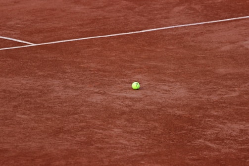A tennis ball rests on a clay tennis court, with visible white boundary lines intersecting across the ground. The court surface has a distinct reddish-brown color, typical of clay courts.