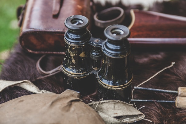 A pair of vintage black binoculars with worn paint rests on a dark furry surface, accompanied by a leather carrying case and part of a rifle. The scene is outdoors, with bits of greenery visible in the blurred background.