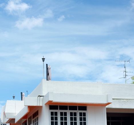 A modern, minimalist white building with large windows and a flat roof under a bright blue sky. Antennas and street lamps protrude from the roof, and there are hints of orange at the building's edges.