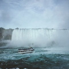 A large waterfall cascades with immense force, creating mist that rises into the air. Below, a tour boat filled with passengers sails through choppy waters close to the waterfall, offering an up-close view of the natural wonder. The scene is enveloped in a cloudy sky, adding to the dramatic effect of the rushing water.