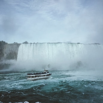 A large waterfall cascades with immense force, creating mist that rises into the air. Below, a tour boat filled with passengers sails through choppy waters close to the waterfall, offering an up-close view of the natural wonder. The scene is enveloped in a cloudy sky, adding to the dramatic effect of the rushing water.