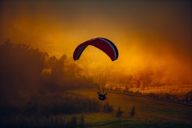 A paraglider floats serenely in the sky against a stunning backdrop of a fiery sunset. The bright orange and yellow hues of the sky illuminate the silhouette of the paraglider, creating a dramatic contrast. Below, the landscape features patches of greenery and clustered trees, adding depth to the scene.