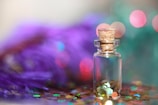 A small glass bottle with a cork stopper containing colorful hexagonal glitter, set against a blurred background. The scene includes vivid colors like purple and teal with bokeh lighting effects.