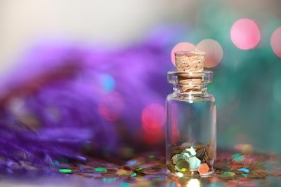 A small glass bottle with a cork stopper containing colorful hexagonal glitter, set against a blurred background. The scene includes vivid colors like purple and teal with bokeh lighting effects.