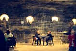 A dimly lit underground dining area with warm pendant lights hanging from the ceiling. The walls have a textured, stone-like appearance with streaks of darker and lighter shades, creating a cozy atmosphere. Several people are seated at wooden tables engaged in conversation, while a server carries a tray with food. The overall setting suggests a casual yet intimate dining experience.
