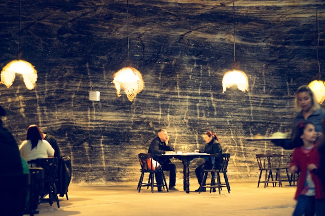 A dimly lit underground dining area with warm pendant lights hanging from the ceiling. The walls have a textured, stone-like appearance with streaks of darker and lighter shades, creating a cozy atmosphere. Several people are seated at wooden tables engaged in conversation, while a server carries a tray with food. The overall setting suggests a casual yet intimate dining experience.