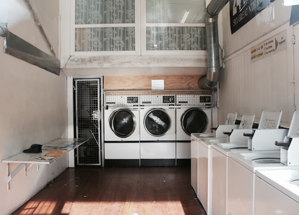 A laundromat interior featuring several washing machines and dryers. The machines are lined up against the walls of a narrow room with smooth wooden floors. Natural light filters through a window above the dryers, casting shadows. There's a small folding table on the left side with a few items on it.