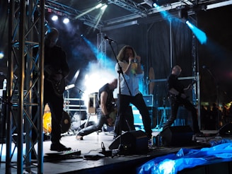 A rock band performs energetically on a stage with elaborate lighting. The lead singer stands at the center, passionately singing into a microphone. Band members are playing guitars, surrounded by a dynamic setup that includes amplifiers and speakers. The scene is filled with smoke effects and dramatic lighting, creating a vibrant and intense atmosphere.