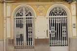 Two ornate, double wrought iron gates with symmetrical designs are set within a pale yellow and beige facade. The gates have intricate scrollwork and are flanked by decorative columns. The building facade shows signs of wear, with chipped paint and an aged appearance. Above the gates, there is a decorative crest resembling wings or a fan.