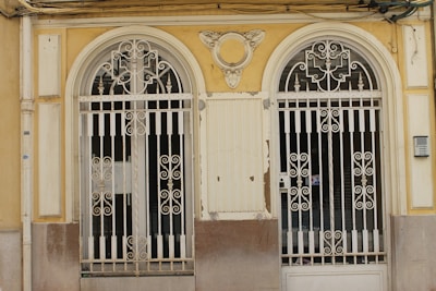 Two ornate, double wrought iron gates with symmetrical designs are set within a pale yellow and beige facade. The gates have intricate scrollwork and are flanked by decorative columns. The building facade shows signs of wear, with chipped paint and an aged appearance. Above the gates, there is a decorative crest resembling wings or a fan.