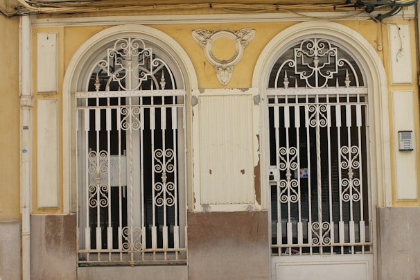 Two ornate, double wrought iron gates with symmetrical designs are set within a pale yellow and beige facade. The gates have intricate scrollwork and are flanked by decorative columns. The building facade shows signs of wear, with chipped paint and an aged appearance. Above the gates, there is a decorative crest resembling wings or a fan.