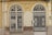 Two ornate, double wrought iron gates with symmetrical designs are set within a pale yellow and beige facade. The gates have intricate scrollwork and are flanked by decorative columns. The building facade shows signs of wear, with chipped paint and an aged appearance. Above the gates, there is a decorative crest resembling wings or a fan.