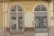 Two ornate, double wrought iron gates with symmetrical designs are set within a pale yellow and beige facade. The gates have intricate scrollwork and are flanked by decorative columns. The building facade shows signs of wear, with chipped paint and an aged appearance. Above the gates, there is a decorative crest resembling wings or a fan.