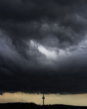 Dark, ominous clouds dominate the sky, creating a dramatic and foreboding atmosphere. A solitary communication tower stands in the distance against a backdrop of darkened landscape, with a faint light on the horizon hinting at an impending storm.
