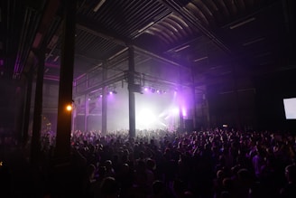 A large crowd of people gathered in a dimly lit industrial-style venue, with high ceilings and visible metal beams. The scene is illuminated by purple lights, creating a vibrant atmosphere as people enjoy what appears to be a concert or music event.