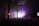 A large crowd of people gathered in a dimly lit industrial-style venue, with high ceilings and visible metal beams. The scene is illuminated by purple lights, creating a vibrant atmosphere as people enjoy what appears to be a concert or music event.