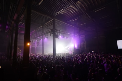A large crowd of people gathered in a dimly lit industrial-style venue, with high ceilings and visible metal beams. The scene is illuminated by purple lights, creating a vibrant atmosphere as people enjoy what appears to be a concert or music event.