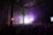 A large crowd of people gathered in a dimly lit industrial-style venue, with high ceilings and visible metal beams. The scene is illuminated by purple lights, creating a vibrant atmosphere as people enjoy what appears to be a concert or music event.