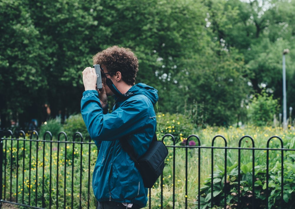 man wearing blue jacket standing and using camera beside flower field surrounded with tall and green trees