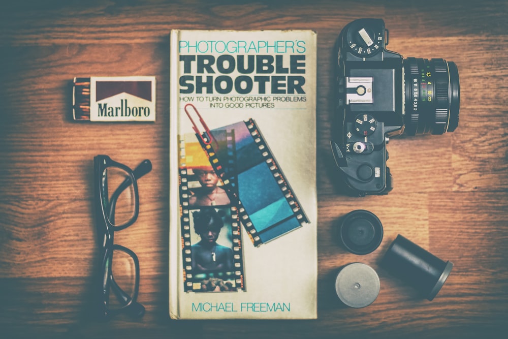 Fotografo Trouble Shooter libro tra fotocamera DSLR, scatola di fiammiferi Marlboro e occhiali da vista