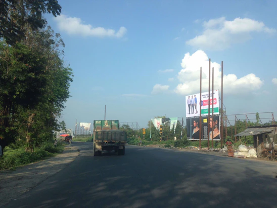 A Nakul Logistics truck on the road with a clear blue sky and lush green fields in the background.