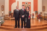 Three people dressed formally stand together in a church setting with wooden flooring and a decorated altar in the background. Red floral arrangements and religious symbols are visible on the walls.