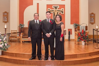 Three people dressed formally stand together in a church setting with wooden flooring and a decorated altar in the background. Red floral arrangements and religious symbols are visible on the walls.