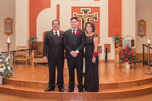 Three people dressed formally stand together in a church setting with wooden flooring and a decorated altar in the background. Red floral arrangements and religious symbols are visible on the walls.