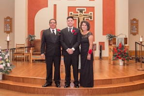 Three people dressed formally stand together in a church setting with wooden flooring and a decorated altar in the background. Red floral arrangements and religious symbols are visible on the walls.
