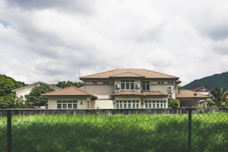 A large, modern house with a beige exterior and brown tiled roof is set against a backdrop of lush green hills and a cloudy sky. The house features multiple windows and air conditioning units. In the foreground, a well-maintained lawn and a metal fence are visible.