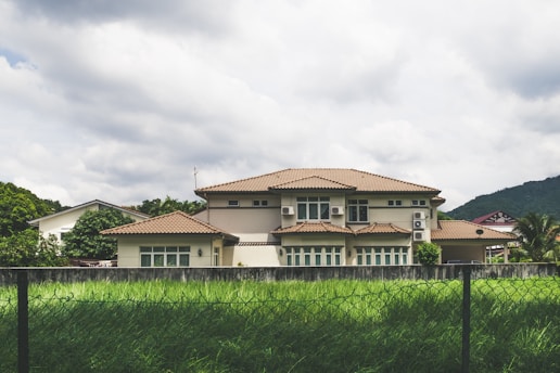 A large, modern house with a beige exterior and brown tiled roof is set against a backdrop of lush green hills and a cloudy sky. The house features multiple windows and air conditioning units. In the foreground, a well-maintained lawn and a metal fence are visible.