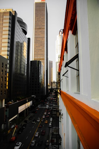 A bustling cityscape with tall modern skyscrapers lining both sides of a street filled with traffic. The buildings have a sleek, glassy finish, reflecting the light from the sky. One of the buildings is distinctively white with orange details, adding a pop of color to the scene.