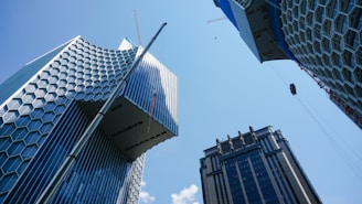 Tall modern skyscrapers with unique architectural designs rise against a clear blue sky. The buildings feature a mix of geometric patterns, such as hexagonal and vertical stripe designs, with cranes visible, indicating ongoing construction.