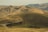 Rolling hills covered with dry grass extend into the distance under a clear sky. A winding road cuts through the landscape, and a herd of cattle is gathered near the foreground. The earthy tones of the hills are dominant throughout the scene.