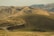 Rolling hills covered with dry grass extend into the distance under a clear sky. A winding road cuts through the landscape, and a herd of cattle is gathered near the foreground. The earthy tones of the hills are dominant throughout the scene.