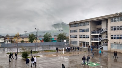 A large, multi-story school building is shown under an overcast sky. There are many people, likely students, standing or walking in the wet courtyard. A foggy hill is visible in the background, and several trees and small buildings surround the area.