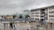 A large, multi-story school building is shown under an overcast sky. There are many people, likely students, standing or walking in the wet courtyard. A foggy hill is visible in the background, and several trees and small buildings surround the area.