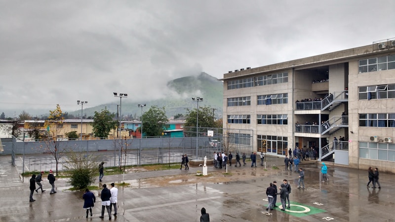 A large, multi-story school building is shown under an overcast sky. There are many people, likely students, standing or walking in the wet courtyard. A foggy hill is visible in the background, and several trees and small buildings surround the area.