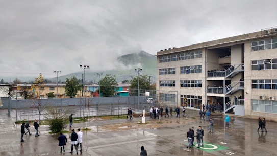 A large, multi-story school building is shown under an overcast sky. There are many people, likely students, standing or walking in the wet courtyard. A foggy hill is visible in the background, and several trees and small buildings surround the area.