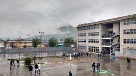 A large, multi-story school building is shown under an overcast sky. There are many people, likely students, standing or walking in the wet courtyard. A foggy hill is visible in the background, and several trees and small buildings surround the area.