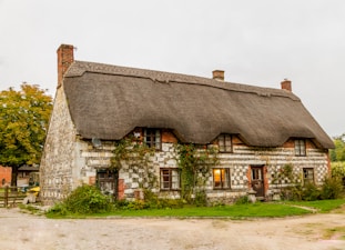 A picturesque, traditional English thatched cottage with stone walls, ivy growing around the windows, and a lush green lawn in the front. The cottage has a rustic charm, with visible brickwork and small windows, one of which is softly illuminated from the inside. The setting appears rural with a gravel path and trees surrounding the area.