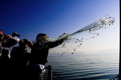 A person is throwing a fishing net into a body of water from a boat or pier, with several people around watching and possibly taking photos. The water is calm and reflects the clear blue sky above.