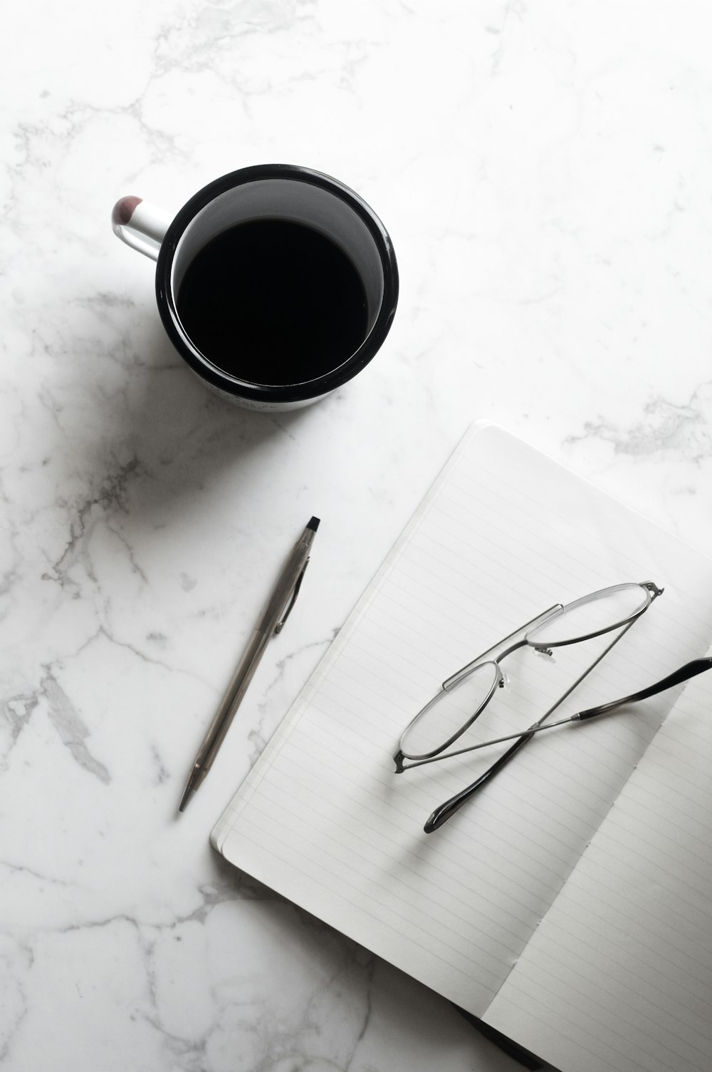 Une tasse de café noir à côté d’un cahier avec un crayon et une paire de lunettes.