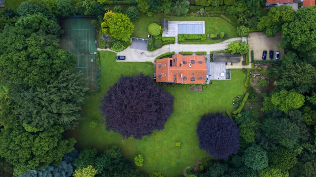 An aerial view of a large estate surrounded by lush greenery and trees. The property features a red-roofed house, a manicured lawn, a driveway with several cars parked, and a tennis court. The estate is surrounded by a dense forest, providing a secluded and tranquil atmosphere.