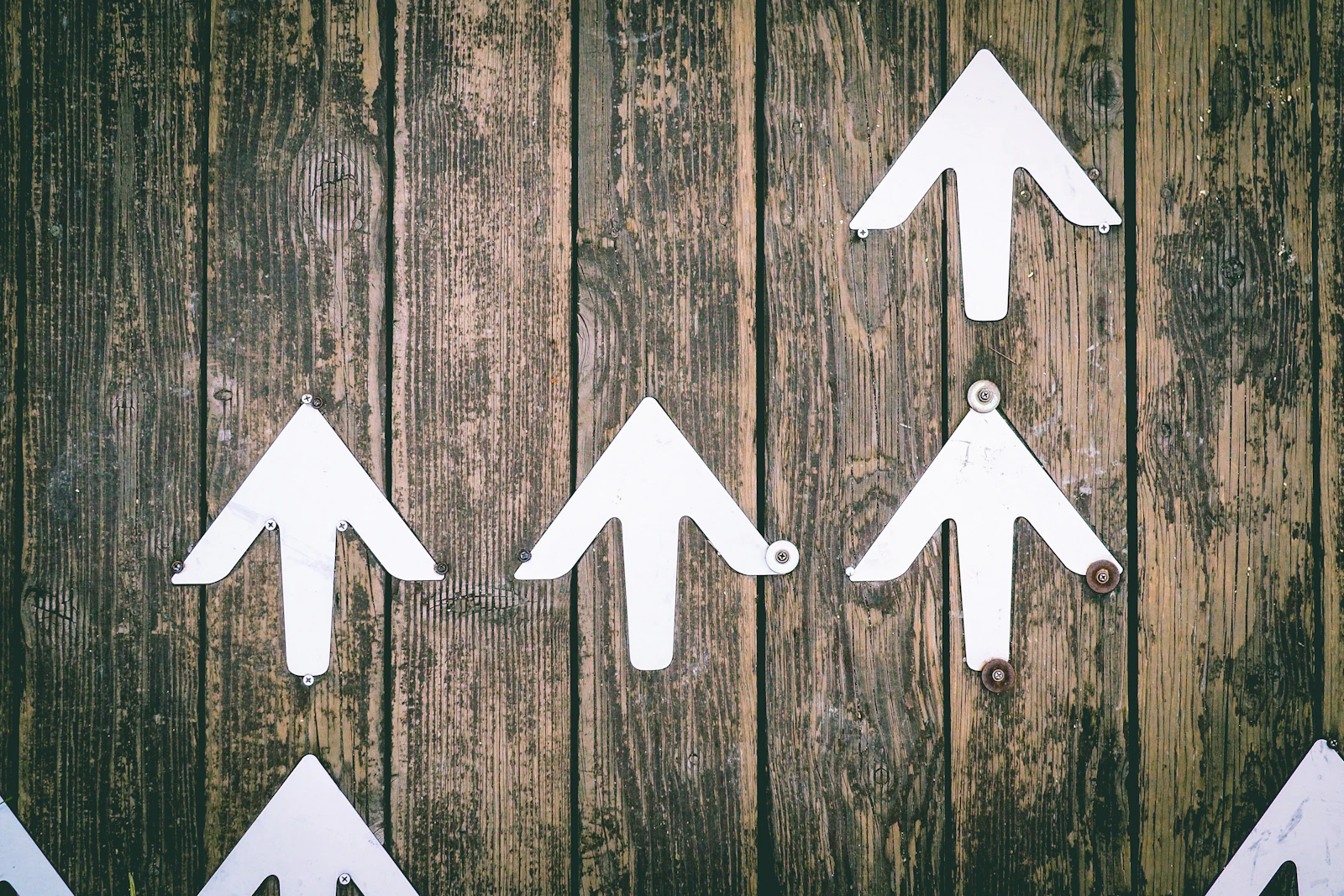 Several white metal arrow signs fixed at different heights on a weathered wooden wall, all pointing upward.